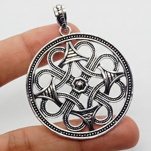 May include: A circular silver-toned metal pendant featuring an intricate openwork design of interlocking loops and geometric patterns. The piece has a polished finish with darkened recessed areas to highlight the detailed metalwork and a top bail.