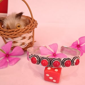 May include: A silver cuff bracelet featuring six red gemstones. The bracelet is displayed on a pink surface with pink flowers and a red die. A small wicker basket with a stuffed animal is in the background.