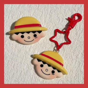 Può includere: Due ciondoli a forma di viso di personaggio dei cartoni animati gialli e rossi, uno con un portachiavi rosso a forma di stella. I ciondoli hanno un cappello giallo con una fascia rossa, capelli neri e un viso sorridente. Lo sfondo è bianco.