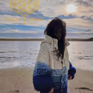 Puede incluir: Un cárdigan de punto grueso con capucha que presenta un degradado de color que va desde el crema en la parte superior hasta el azul claro, azul medio y azul marino oscuro en el dobladillo. Incluye cierre de botones y el texto NÚCLEO ALQUIMIA.