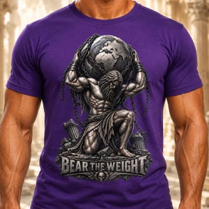 Könnte beinhalten: Ein violettes Kurzarm-T-Shirt mit einem Grafikdruck einer muskulösen, bärtigen Figur, die kniet und einen großen Globus auf den Schultern trägt, umwickelt mit schweren Ketten. Der Text "BEAR THE WEIGHT" ist darunter in Metallschrift gedruckt.