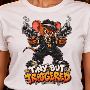 May include: A white t-shirt featuring a cartoon mouse dressed in a black pinstripe suit, fedora, and gold chain, holding two smoking revolvers. Below the mouse, the text reads "TINY BUT TRIGGERED" in bold, orange, and white graffiti-style lettering.