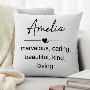 Puede incluir: Cojín blanco con el nombre "Amelia" en escritura negra, un icono de corazón y las palabras "marvelous, caring, beautiful, kind, loving" en negro. El cojín está sobre un sofá gris.