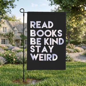 May include: A black garden flag with the words "READ BOOKS BE KIND STAY WEIRD" in white, displayed in a garden setting. The flag is mounted on a black metal pole, with a house and garden in the background.
