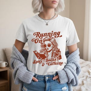 Può includere: T-shirt bianco con grafica marrone che recita "Running On Spicy Books & Caffeine". La grafica presenta una donna con occhiali da sole, che legge un libro e tiene in mano una bevanda al caffè. La maglietta è in stile casual.