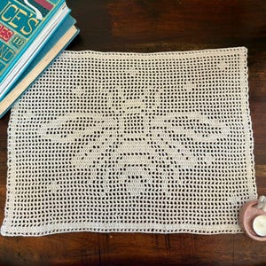 Könnte beinhalten: Cremefarbene, rechteckige Häkeldecke mit einem zentralen Sternmuster. Das durchbrochene Muster ist über das gesamte Stück sichtbar. Ein kleiner, rosa Keramik-Kerzenhalter mit einer weißen Figur und einer brennenden Kerze befindet sich rechts. Bücher sind oben links zu sehen.