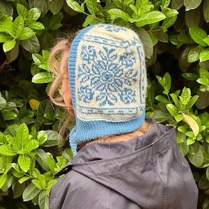 Peut inclure: Un bonnet tricoté bleu et crème avec un motif flocon de neige. Le bonnet présente une base tricotée bleu clair avec un panneau carré crème sur le dessus, orné d'un motif de flocon de neige bleu détaillé. Le bonnet est conçu pour couvrir la tête et le cou.