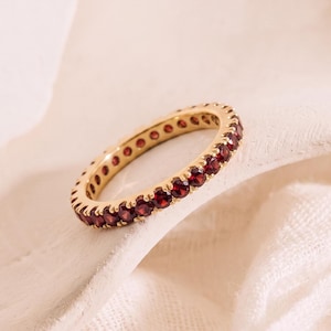 Könnte beinhalten: Ein goldener Eternity-Ring mit einer durchgehenden Reihe kleiner, runder, tiefroter Granat-Edelsteine, die in einen polierten Metallrahmen gefasst sind. Der Ring ist kreisförmig mit gleichbleibender Breite und einer einheitlichen Anordnung der Steine.