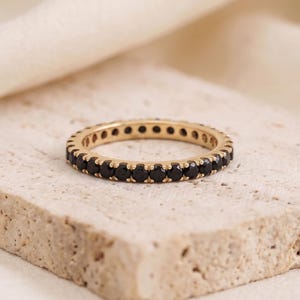 Puede incluir: Un anillo de eternidad en oro con una hilera continua de pequeñas piedras preciosas redondas de color negro engastadas en garras. El anillo tiene un acabado metálico pulido y una forma circular uniforme con piedras oscuras alrededor.