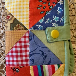 May include: A rectangular fabric cover featuring a patchwork design of various colourful patterns including yellow gingham, red and white checks, floral prints, and stripes. The cover is edged in light green fabric and has a yellow snap button closure.