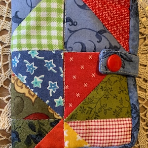 May include: A rectangular quilted fabric case featuring a patchwork design of various patterns including green and white gingham, red and white check, floral prints, and solid colours. The case has a blue border and a single bright red snap button closure.