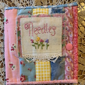 May include: A square fabric needle case featuring a patchwork design of pink, blue, and yellow floral and gingham patterns. A central cream-coloured fabric patch is embroidered with the word Needles and small flowers, finished with a pink snap button.