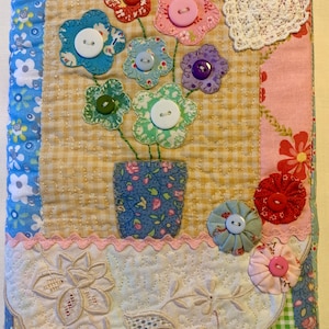 May include: A rectangular fabric book cover featuring a quilted patchwork design with floral patterns in blue, pink, yellow, and green. The centre displays a fabric vase with appliqued flowers made of patterned fabric and colourful buttons.