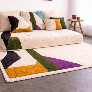 Op de afbeelding: Een crèmekleurig tapijt en een bijpassende bank met geometrische ontwerpen in zwart, groen, geel en paars. De bank heeft verschillende sierkussens in diverse kleuren. Twee kleine houten bijzettafels zijn zichtbaar.