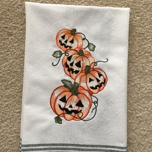 May include: White kitchen towel with an embroidered design of orange jack-o'-lanterns with black faces and green vines. The bottom of the towel has a gray stripe design.
