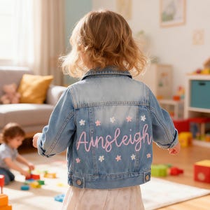 May include: Pale blue denim jacket featuring the name "Ainsleigh" in pink script, decorated with small white and pink flower motifs. The jacket is designed for a child.
