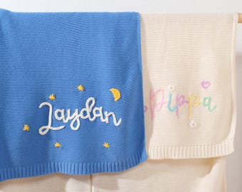Puede incluir: Dos mantas personalizadas para bebés. Una es azul con el nombre "Jaydan" bordado en blanco, junto con estrellas amarillas y una luna creciente. La otra es de color crema con el nombre "Pippa" bordado en colores pastel, con un corazón y una flor.