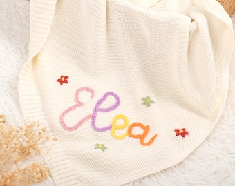May include: Cream-coloured knitted baby blanket with the name "Elea" embroidered in colourful thread. The blanket features small embroidered flowers in various colours. The blanket is folded and placed on a white, fluffy surface.