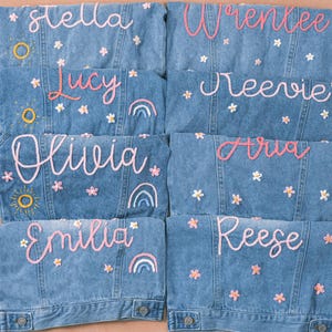 May include: A collection of personalized denim jackets in various shades of blue. Each jacket features a name embroidered in pink script, along with floral and rainbow embellishments. The jackets are arranged on a neutral background.