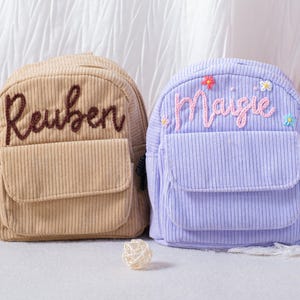 Könnte beinhalten: Zwei Cord-Rucksäcke, einer in Beige und einer in Lavendel, werden gezeigt. Der beige Rucksack hat den Namen "Reuben" in Braun gestickt, während der lavendelfarbene Rucksack den Namen "Maisie" in Rosa mit floralen Akzenten hat. Beide haben eine Vordertasche.