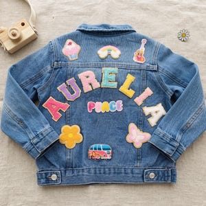 May include: A blue denim jacket featuring the name AURELIA in colorful fabric letters across the back. The jacket is decorated with various embroidered patches including a cupcake, rainbow, guitar, flower, butterfly, van, and the word PEACE.