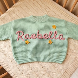 Puede incluir: Un suéter de punto de manga larga en color verde menta con el nombre Raebella bordado en hilo rosa en el centro. El suéter está decorado con tres pequeñas aplicaciones de flores tridimensionales amarillas con centros rosas.