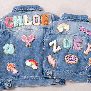 Könnte beinhalten: Zwei hellblaue Jeansjacken mit bunten gestickten Aufnähern. Eine Jacke trägt den Schriftzug "CHLOE" und die andere "ZOE". Zu den Aufnähern gehören ein Regenbogen, Herzen, ein Donut und Tennisschläger.
