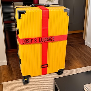 Può includere: Una valigia gialla con una cinghia rossa con la scritta "JOSH'S LUGGAGE". La valigia ha protezioni angolari nere ed è su un tappeto chiaro. La stanza ha pavimenti in legno. La valigia è su ruote.