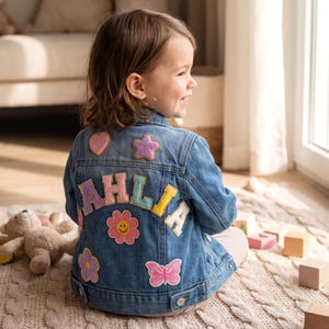 Puede incluir: Una chaqueta de mezclilla azul con el nombre DAHLIA en letras grandes, coloridas y texturizadas en la parte posterior. La chaqueta está decorada con varios parches, incluyendo un corazón rosa, una estrella morada, una flor rosa con cara sonriente, una flor morada y una mariposa rosa.