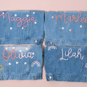 May include: Four light blue denim jackets with personalized names embroidered in pink. Each jacket features floral and rainbow embellishments. The names are Maggie, Marlee, Olivia, and Lilah.