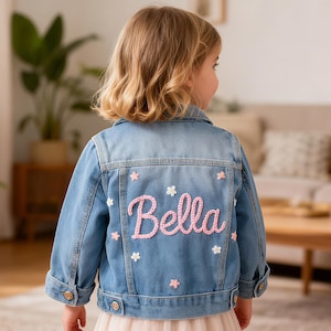 May include: A light blue denim jacket with the name "Bella" in pink script lettering on the back. The jacket is decorated with small pink and white flower appliqués. The jacket has button closures and pockets.