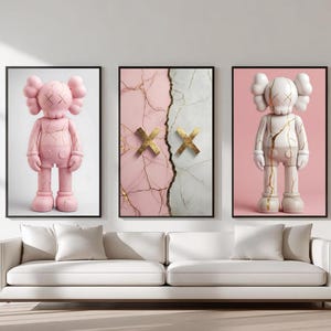 May include: Three framed art prints. One features a pink cartoon figure, another a pink and white marble design with gold detailing, and the third a white cartoon figure. The prints are displayed above a white sofa.