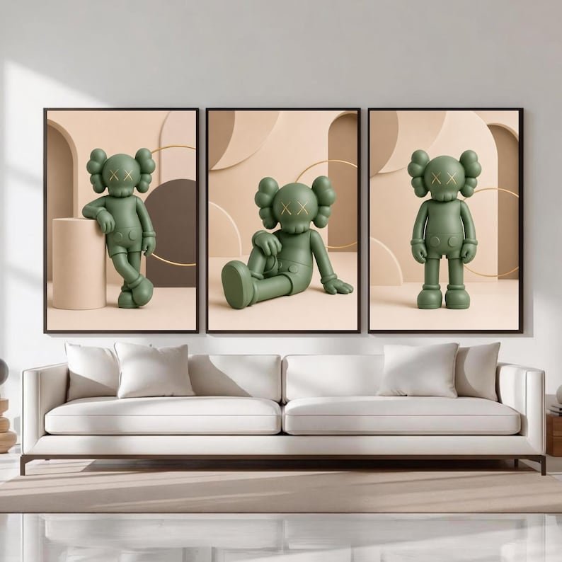 May include: A set of three framed wall art prints featuring a matte olive green cartoon character with X-shaped eyes. Each print displays the character in a different pose against a neutral beige background with geometric shapes and thin gold rings.
