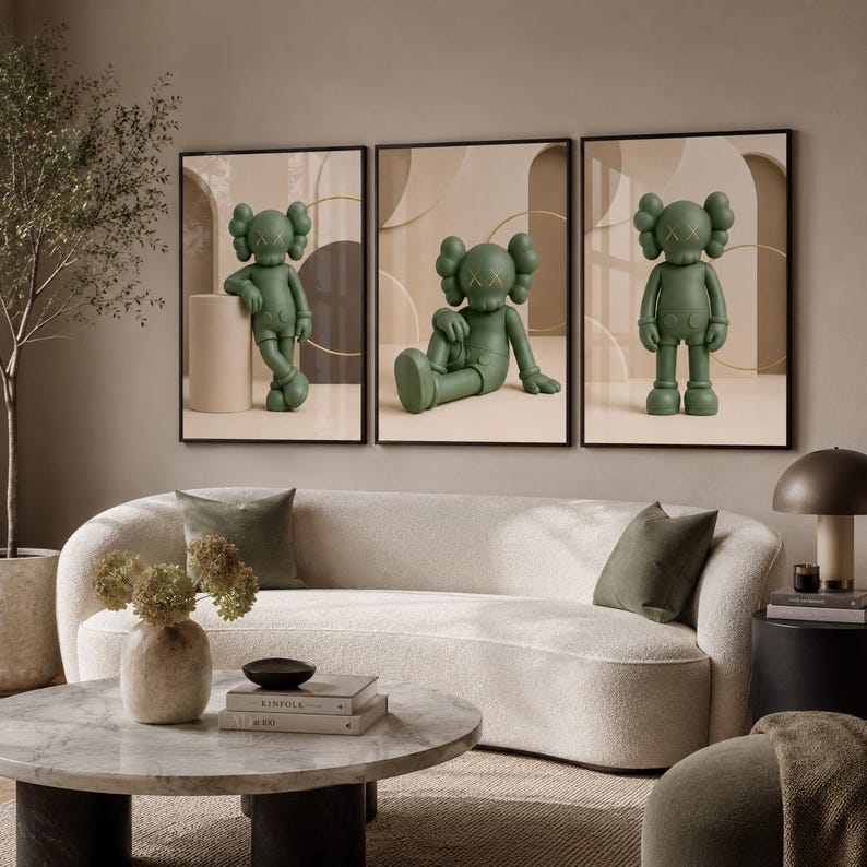 May include: A set of three framed wall art prints featuring a matte olive green cartoon character with X-shaped eyes. Each print displays the character in a different pose against a neutral beige background with abstract geometric shapes and arches.
