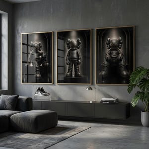 May include: Three framed wall art prints featuring a black character with X-shaped eyes and ears, depicted in different poses within a dark, arched setting. The prints are displayed in thin gold frames on a grey wall above a long black console table.