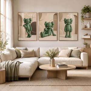 May include: A set of three framed wall art prints featuring a matte olive green cartoon character with X-shaped eyes. Each print displays the character in a different pose against a neutral beige background with abstract geometric shapes and circles.