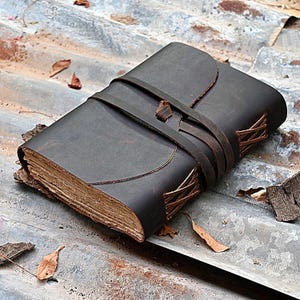May include: A dark brown leather journal with a wrap-around strap closure. The cover features a smooth, matte finish with visible stitching along the spine edges. The pages are thick, textured, and cream-colored, showing a deckled edge profile.