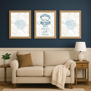 May include: Three framed wall art prints with a coastal theme. Each print features a light blue seashell design on a white and gray wavy background. The center print has a pearl design and the text "Shine Quietly Like A Pearl."