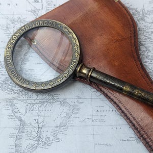 May include: Antique brass magnifying glass with the text "HENRY HUGHES SON LTD. LONDON 1941" around the rim. The magnifying glass rests on a vintage map and a brown leather case. The handle has an ornate floral design.