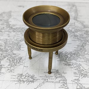 May include: A vintage brass magnifying glass with a round lens and a three-legged stand. The base of the stand is inscribed with "ROSS LONDON". The magnifying glass is set against a backdrop of a world map.