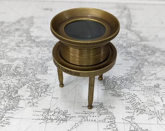 May include: A vintage brass magnifying glass with a round lens and a three-legged stand. The base of the stand is inscribed with "ROSS LONDON". The magnifying glass is set against a backdrop of a world map.