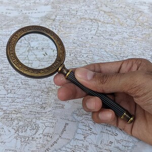 May include: A vintage brass magnifying glass with a black and gold handle, held over a detailed map. The magnifying glass is engraved with "HENRY HUGHES SON LTD LONDON - 1941". A classic tool for reading maps or examining small details.