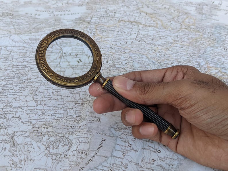 May include: A vintage brass magnifying glass with a black and gold handle, held over a detailed map. The magnifying glass is engraved with "HENRY HUGHES SON LTD LONDON - 1941". A classic tool for reading maps or examining small details.