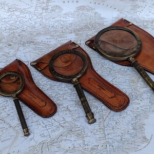 May include: Three antique brass magnifying glasses with dark brown leather cases are displayed on a vintage map. The magnifying glasses have detailed handles and circular lenses. The leather cases are embossed with text.