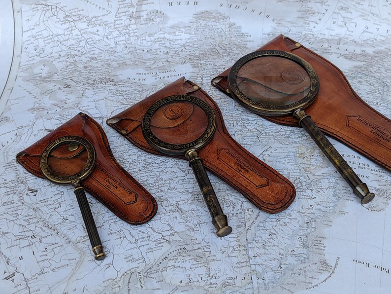 May include: Three antique brass magnifying glasses with dark brown leather cases are displayed on a vintage map. The magnifying glasses have detailed handles and circular lenses. The leather cases are embossed with text.