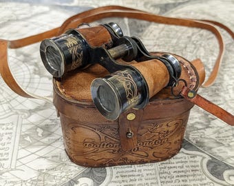 Könnte beinhalten: Antikes Fernglas aus Messing und Leder mit passendem Lederetui. Das Fernglas hat braune Ledergriffe und ist mit floralen Mustern graviert. Auf dem Etui steht "Dollon London". Ein brauner Lederriemen ist befestigt.