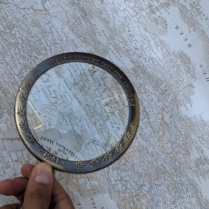 May include: A vintage brass magnifying glass with a clear lens, resting on a detailed antique map. The magnifying glass has the inscription "HENRY HUGHES & SON LTD LONDON" and the year "1941". The map features geographical details.