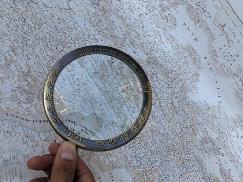 May include: A vintage brass magnifying glass with a clear lens, resting on a detailed antique map. The magnifying glass has the inscription "HENRY HUGHES & SON LTD LONDON" and the year "1941". The map features geographical details.