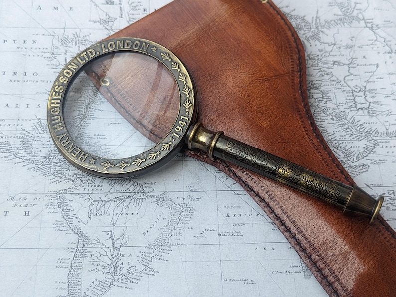 May include: Antique brass magnifying glass with the text "HENRY HUGHES SON LTD. LONDON 1941" around the rim. The magnifying glass rests on a vintage map and a brown leather case. The handle has an ornate floral design.