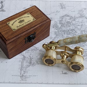 May include: Vintage brass marine binoculars with a mother-of-pearl handle, resting on a map. A wooden box with a brass label that reads "MARINE BINOCULAR, Dallond London" is also in the image.